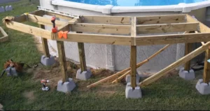 Partially constructed wooden deck framework surrounding an above-ground pool, showing the structural supports and tools used in the building process. Ready to transform your pool area? Learn how to build an above-ground pool deck today!