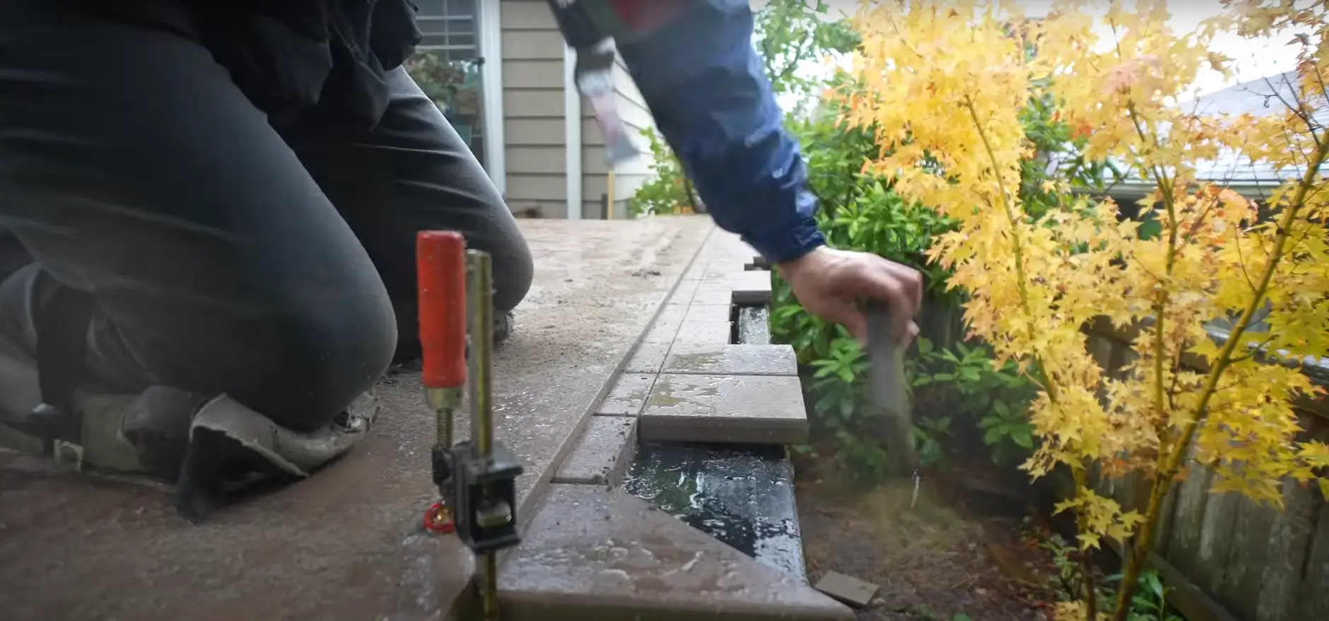 A close-up of a worker using hidden fasteners to secure composite decking boards, surrounded by lush autumn foliage.